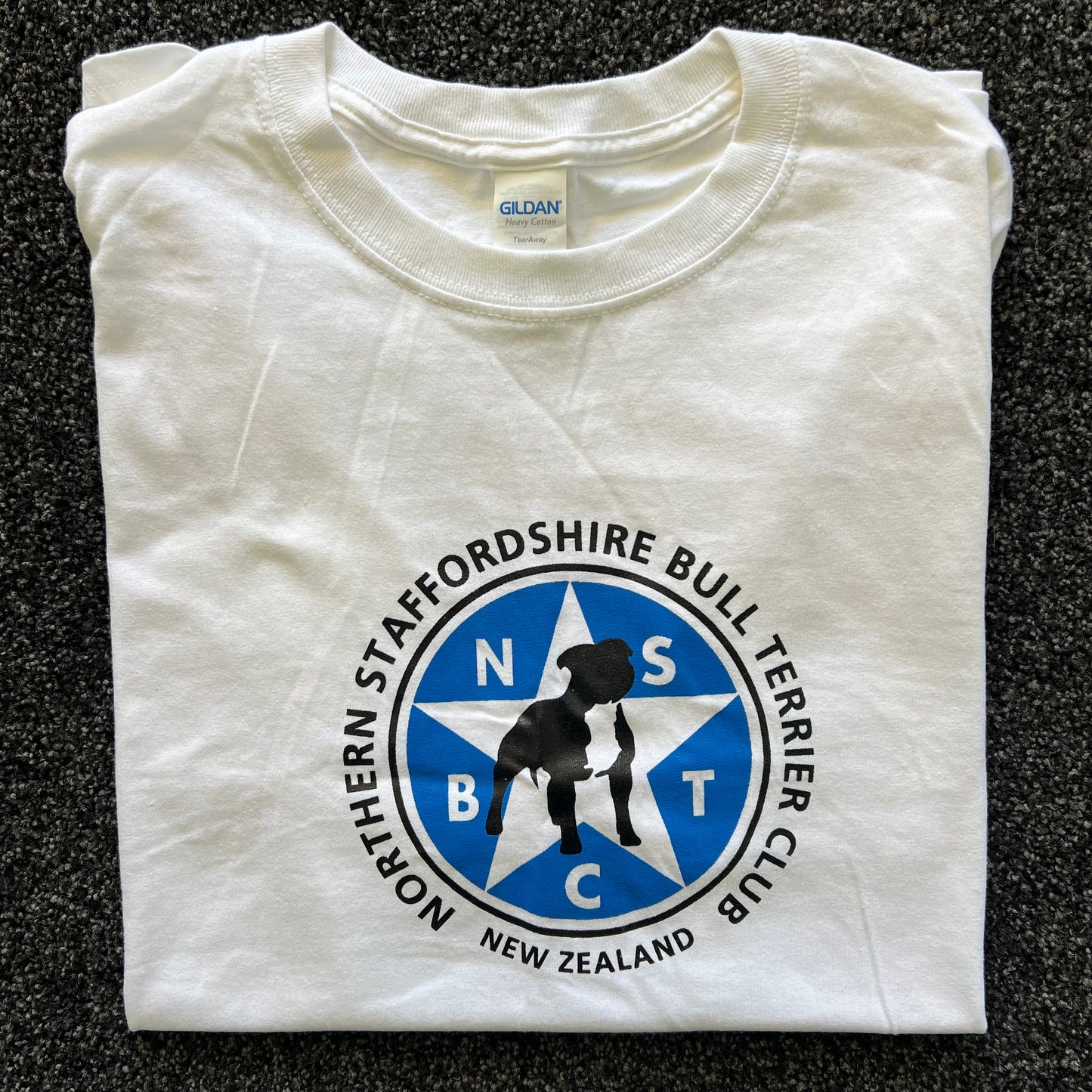 Folded white t-shirt with Northern Staffordshire Bull Terrier Club logo on a dark textured surface